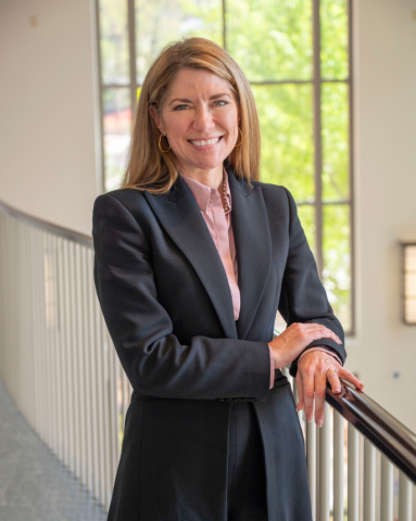 A woman in a black suit smiles confidently, leaning on a railing in a bright room with large windows, natural light streaming in and greenery outside.