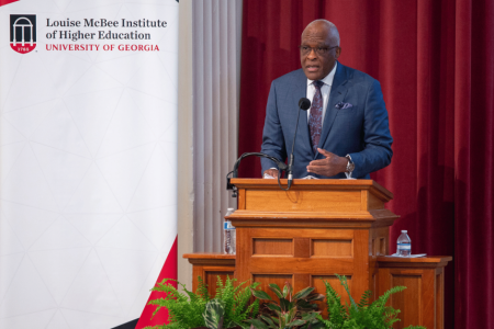 A man in a suit speaks at a wooden podium with a microphone, in front of red curtains and a University of Georgia banner. The setting suggests a formal event.